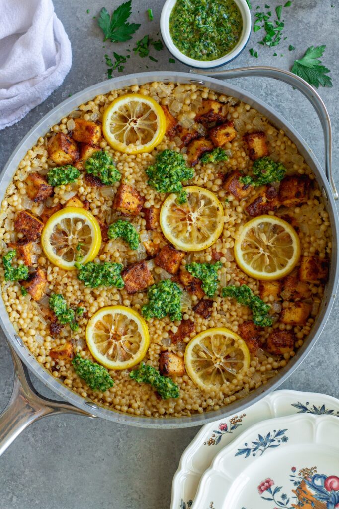 Golden crispy tofu cubes and fluffy lemon couscous in a skillet topped with green chimichurri sauce and lemon slices.