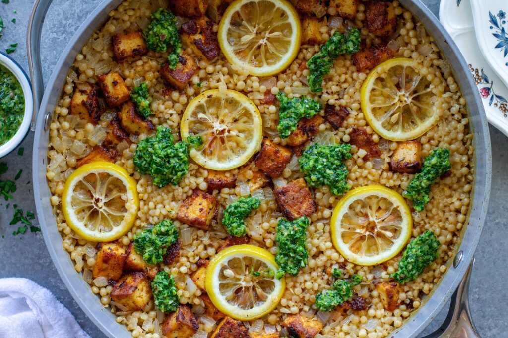Golden crispy tofu cubes and fluffy lemon couscous in a skillet topped with green chimichurri sauce and lemon slices.