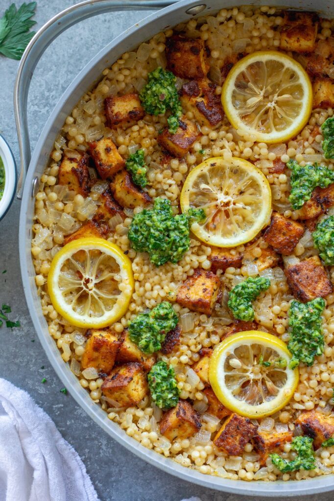 Golden crispy tofu cubes and fluffy lemon couscous in a skillet topped with green chimichurri sauce and lemon slices.