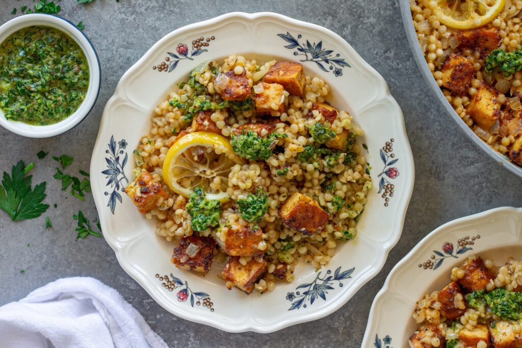 Golden crispy tofu cubes and fluffy lemon couscous in a skillet topped with green chimichurri sauce and lemon slices.