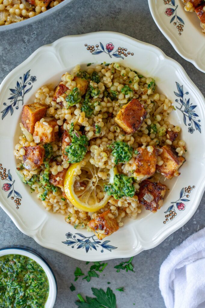 Golden crispy tofu cubes and fluffy lemon couscous in a skillet topped with green chimichurri sauce and lemon slices.
