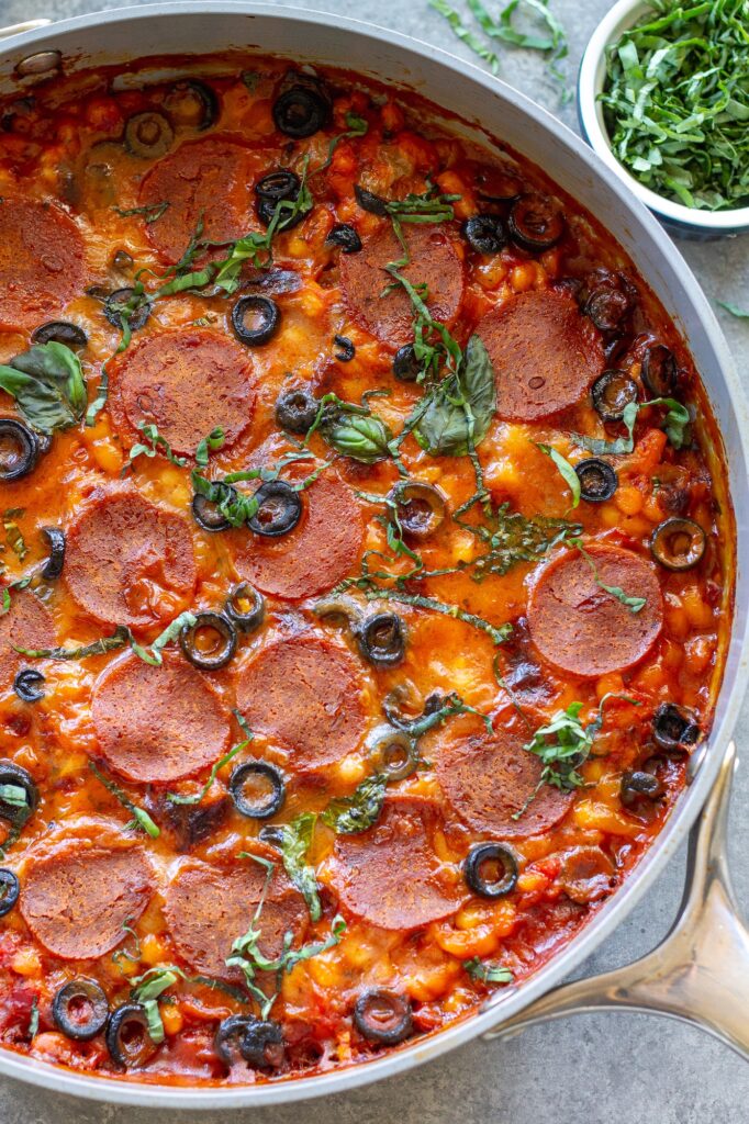 Vegan pizza beans in a skillet topped with melted vegan mozzarella, pepperoni, parmesan and fresh basil