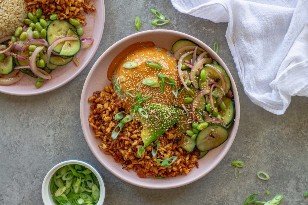 Vegan shredded tofu sushi bowl with sriracha sauce, cucumber edamame salad, avocado, and toasted sesame seeds