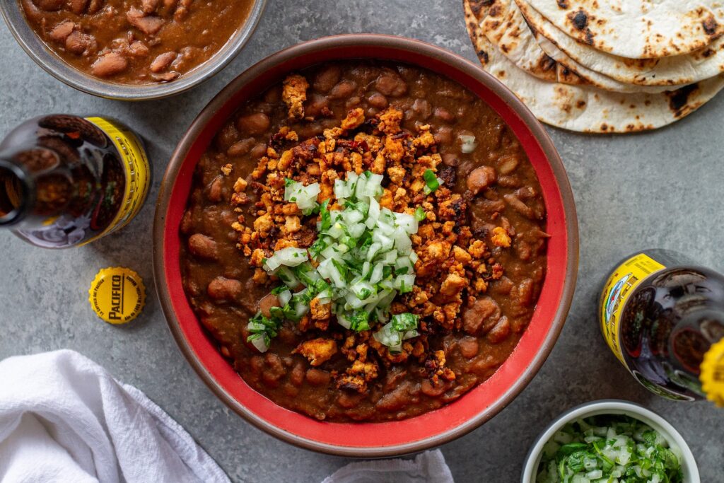 Serving of vegan charro beans topped with smoky tofu crumbles and minced cilantro and onion, with a side of lime wedges and warm tortillas.