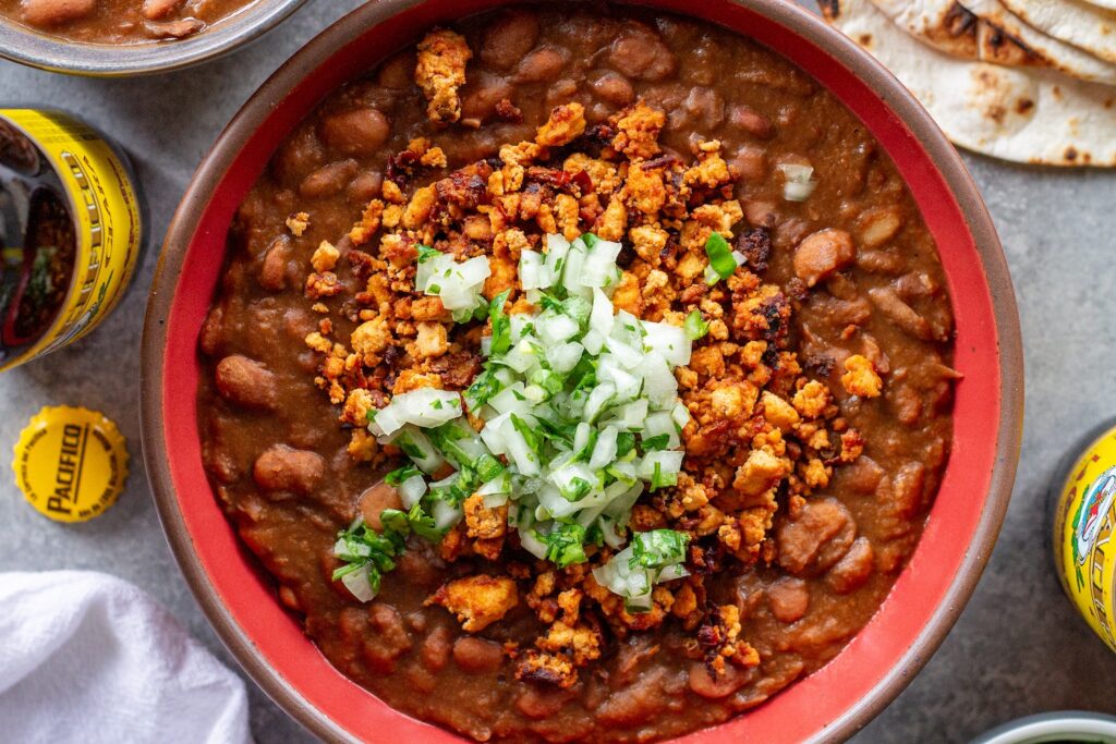 Serving of vegan charro beans topped with smoky tofu crumbles and minced cilantro and onion, with a side of lime wedges and warm tortillas.