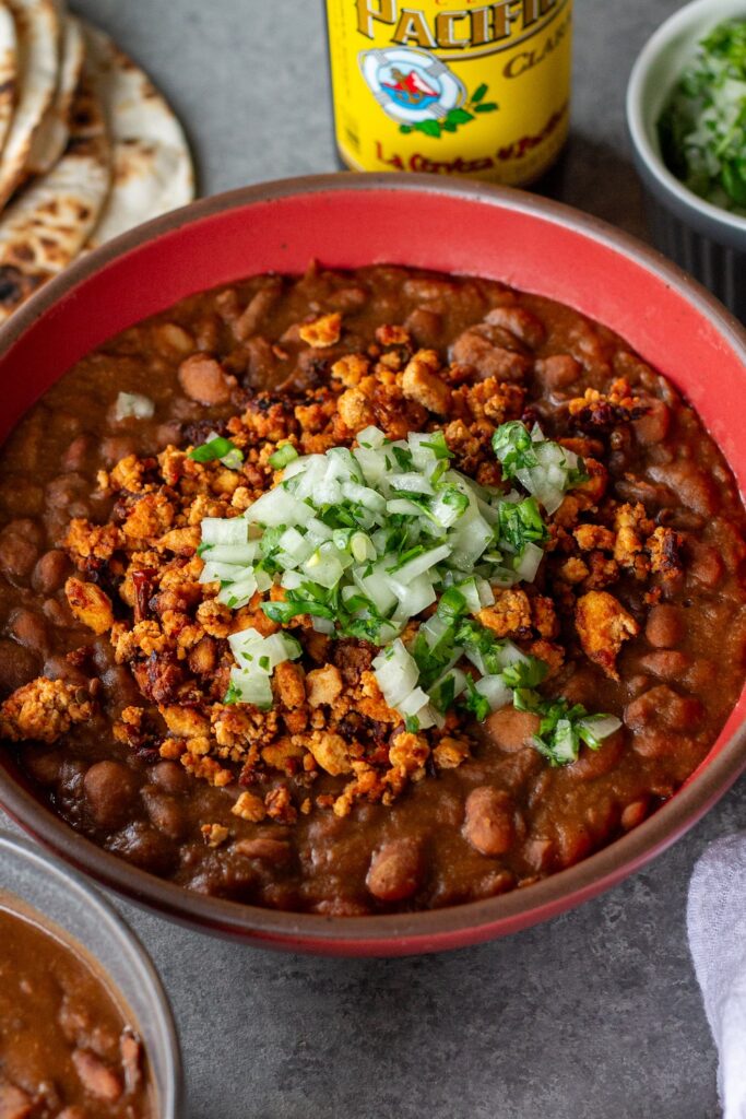 Serving of vegan charro beans topped with smoky tofu crumbles and minced cilantro and onion, with a side of lime wedges and warm tortillas.