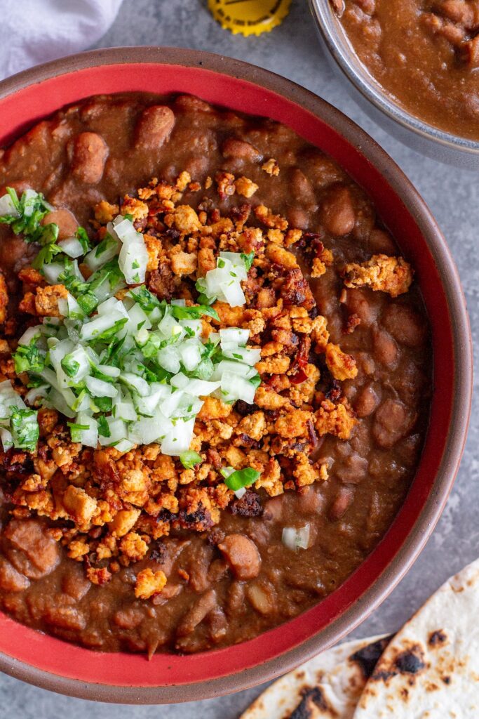 Serving of vegan charro beans topped with smoky tofu crumbles and minced cilantro and onion, with a side of lime wedges and warm tortillas.