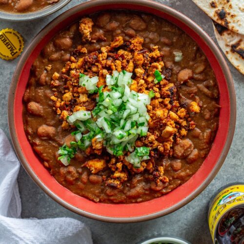 Serving of vegan charro beans topped with smoky tofu crumbles and minced cilantro and onion, with a side of lime wedges and warm tortillas.
