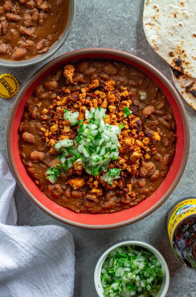 Serving of vegan charro beans topped with smoky tofu crumbles and minced cilantro and onion, with a side of lime wedges and warm tortillas.