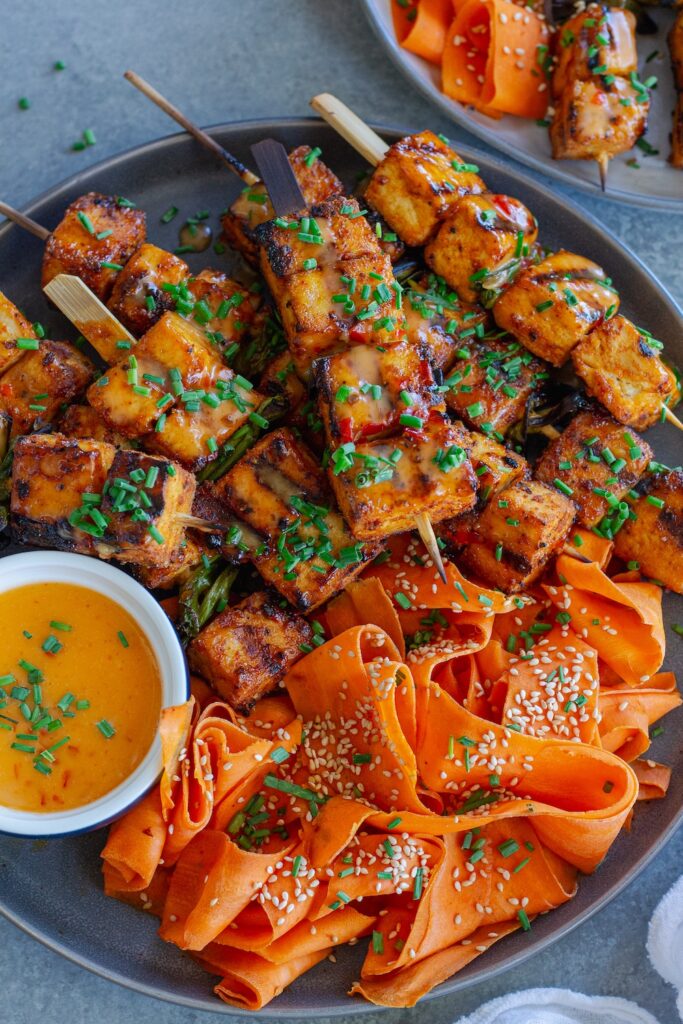 Grilled Bang Bang Tofu Skewers with charred edges and a drizzle of creamy bang bang sauce, garnished with green chives and served next to a refreshing shaved carrot salad.