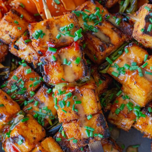 Grilled Bang Bang Tofu Skewers with charred edges and a drizzle of creamy bang bang sauce, garnished with green chives and served next to a refreshing shaved carrot salad.
