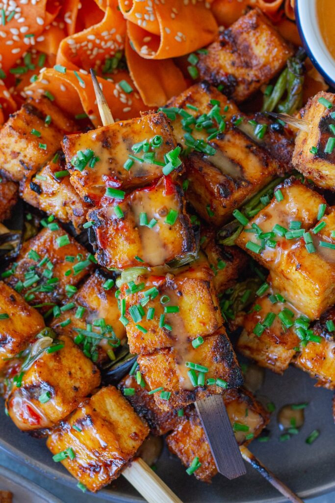 Grilled Bang Bang Tofu Skewers with charred edges and a drizzle of creamy bang bang sauce, garnished with green chives and served next to a refreshing shaved carrot salad.