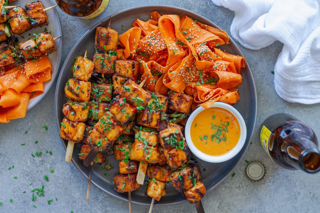 Grilled Bang Bang Tofu Skewers with charred edges and a drizzle of creamy bang bang sauce, garnished with green chives and served next to a refreshing shaved carrot salad.