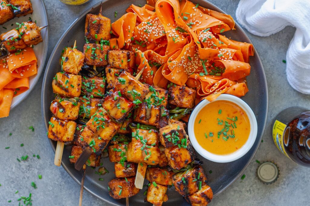 Grilled Bang Bang Tofu Skewers with charred edges and a drizzle of creamy bang bang sauce, garnished with green chives and served next to a refreshing shaved carrot salad.
