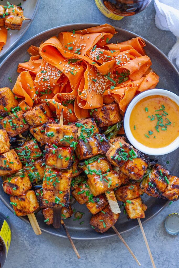 Grilled Bang Bang Tofu Skewers with charred edges and a drizzle of creamy bang bang sauce, garnished with green chives and served next to a refreshing shaved carrot salad.