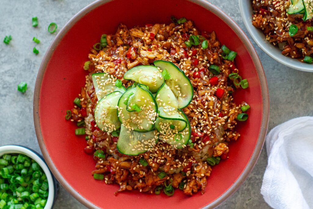 Beijing Beef-Inspired bowl with saucy shredded tofu served over brown rice with vibrant red peppers and garnished with a fresh cucumber salad, sesame seeds and green onion