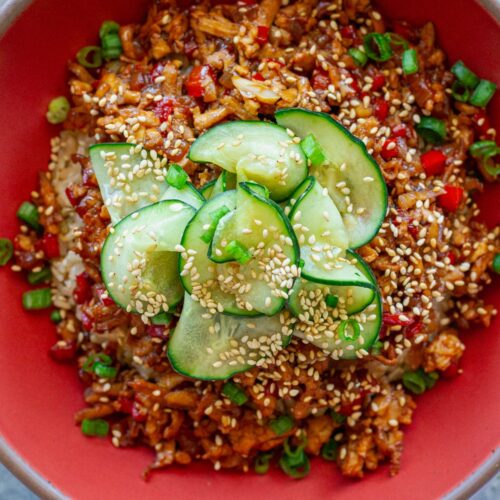 Beijing Beef-Inspired bowl with saucy shredded tofu served over brown rice with vibrant red peppers and garnished with a fresh cucumber salad, sesame seeds and green onion