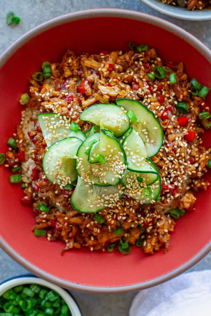Beijing Beef-Inspired bowl with saucy shredded tofu served over brown rice with vibrant red peppers and garnished with a fresh cucumber salad, sesame seeds and green onion