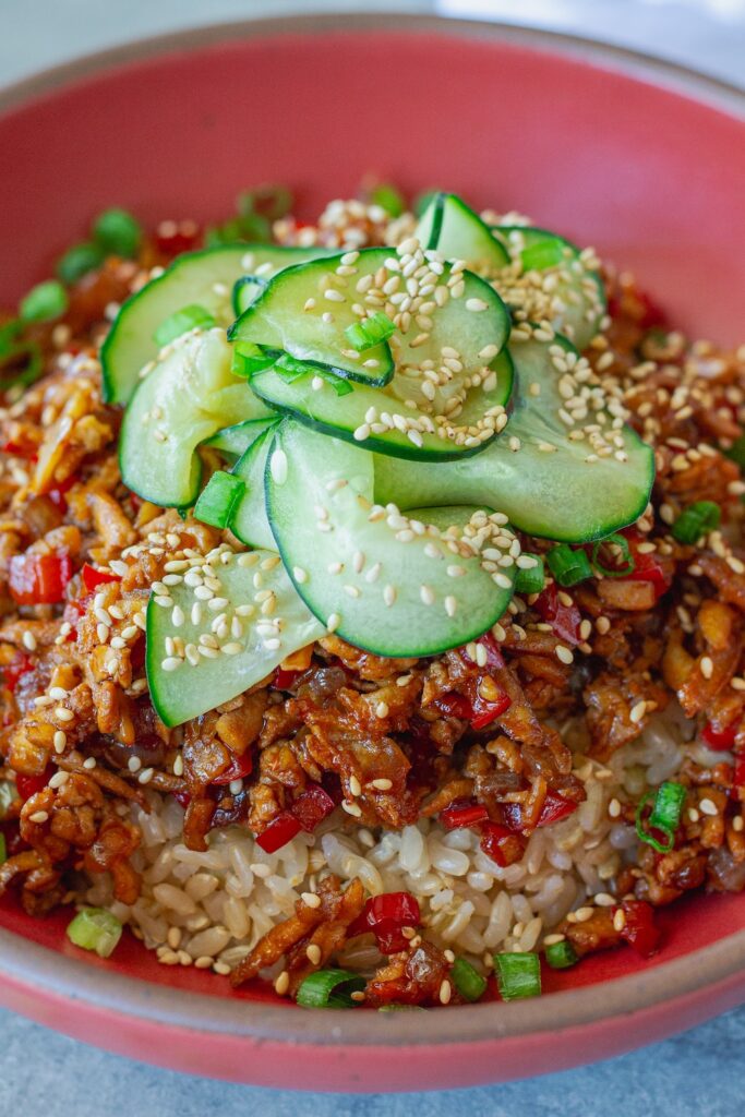 Beijing Beef-Inspired bowl with saucy shredded tofu served over brown rice with vibrant red peppers and garnished with a fresh cucumber salad, sesame seeds and green onion