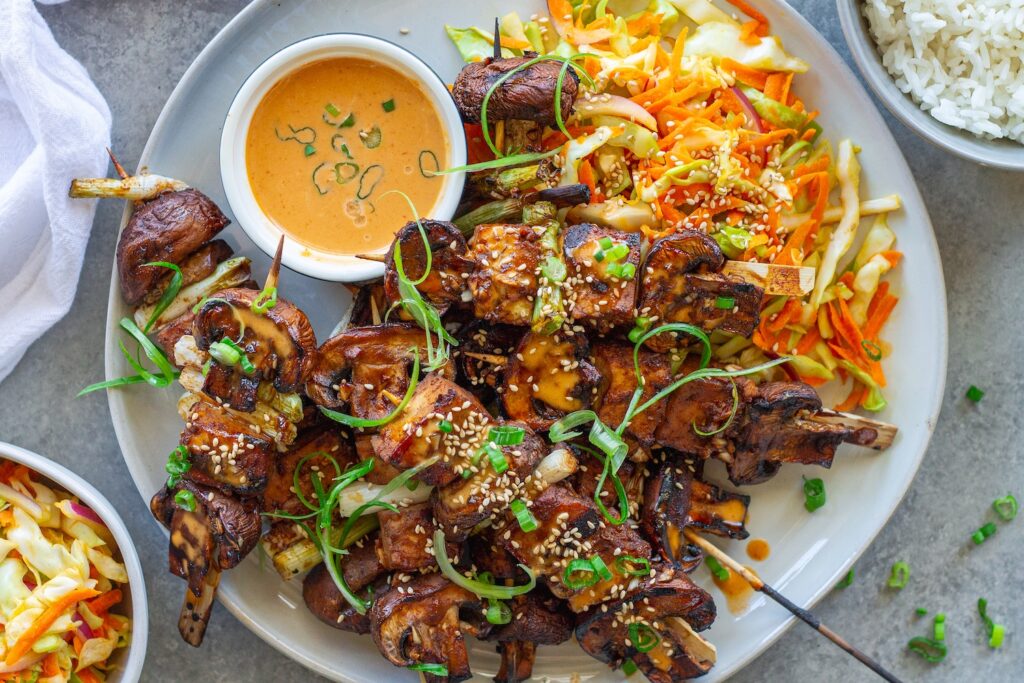 Golden grilled Korean BBQ tofu skewers with mushrooms and scallions on bamboo skewers, served with a side of Gochujang mayonnaise and garnished with sesame seeds and green onions