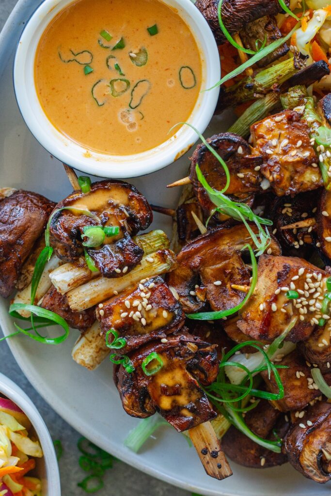 Golden grilled Korean BBQ tofu skewers with mushrooms and scallions on bamboo skewers, served with a side of Gochujang mayonnaise and garnished with sesame seeds and green onions