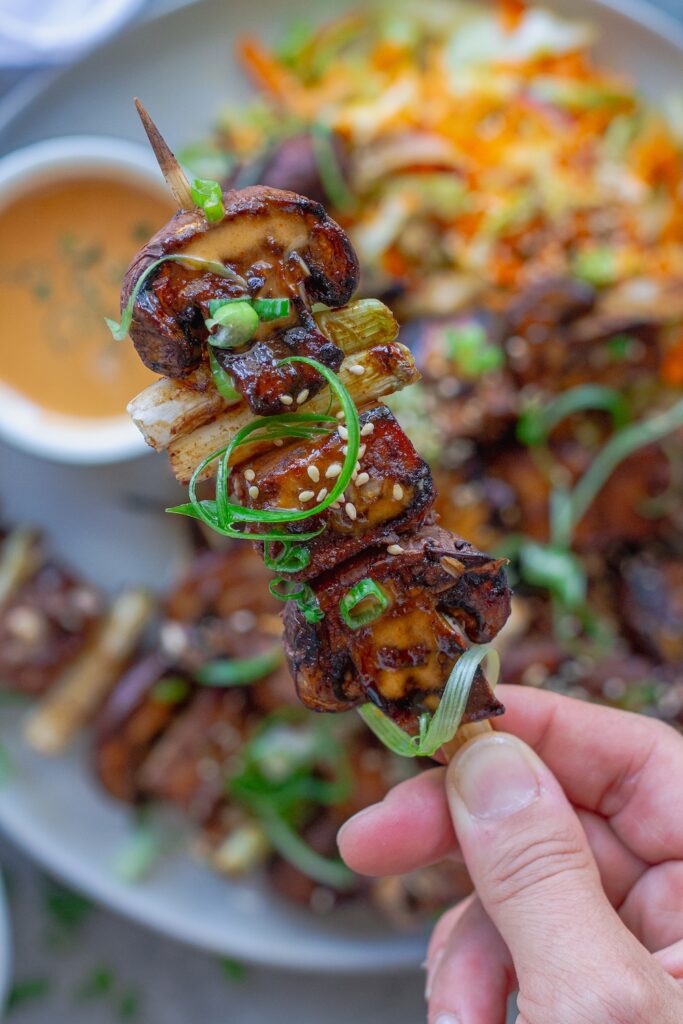 Golden grilled Korean BBQ tofu skewers with mushrooms and scallions on bamboo skewers, served with a side of Gochujang mayonnaise and garnished with sesame seeds and green onions