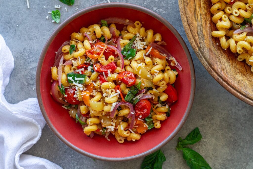 A colorful bowl of roasted sweet pepper pasta salad topped with fresh basil and vegan parmesan.