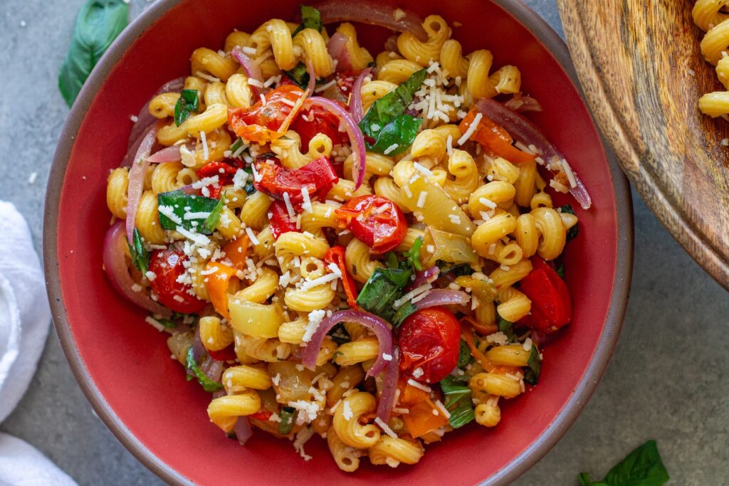 A colorful bowl of roasted sweet pepper pasta salad topped with fresh basil and vegan parmesan.