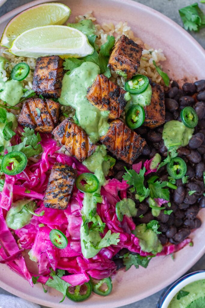 Vegan fish taco bowl with crispy blackened tofu, purple pickled cabbage, cilantro-lime rice, creamy avocado dressing, and garnished with fresh cilantro, jalapeño slices, and a lime wedge