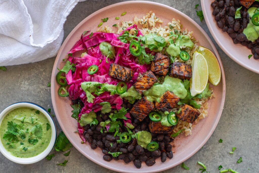 Vegan fish taco bowl with crispy blackened tofu, purple pickled cabbage, cilantro-lime rice, black beans, creamy avocado dressing, and garnished with fresh cilantro, jalapeño slices, and a lime wedge