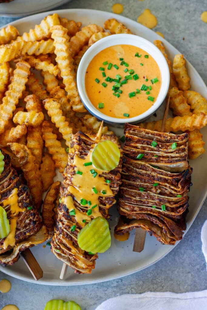 Golden brown vegan cheeseburger tortilla kabobs on wooden skewers with burger sauce drizzle and fresh dill pickles