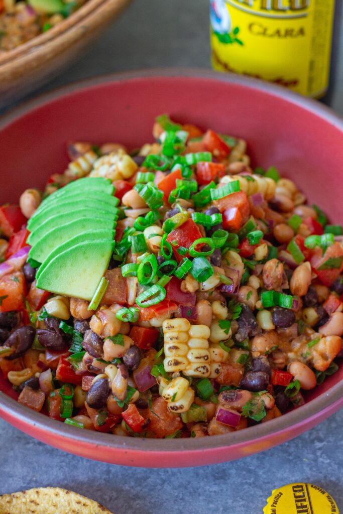 A bowl of colorful cowboy caviar with black beans, corn, diced tomatoes, red bell pepper, and black-eyed peas tossed in a smoky chipotle-lime dressing, garnished with fresh cilantro, served with tortilla chips on the side.
