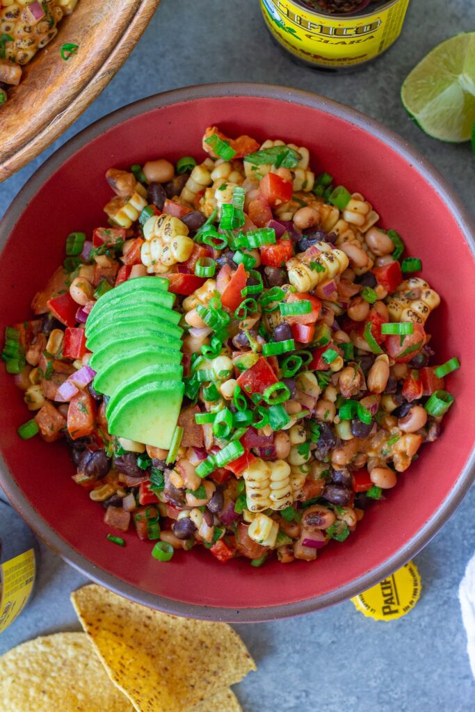A bowl of colorful cowboy caviar with black beans, corn, diced tomatoes, red bell pepper, and black-eyed peas tossed in a smoky chipotle-lime dressing, garnished with fresh cilantro, served with tortilla chips on the side.