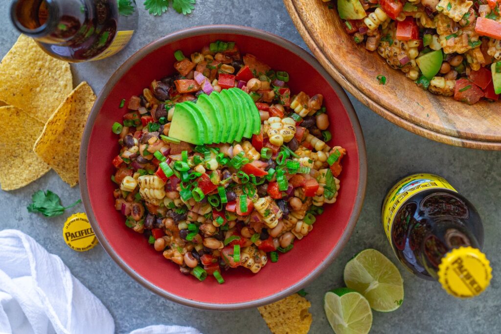 A bowl of colorful cowboy caviar with black beans, corn, diced tomatoes, red bell pepper, and black-eyed peas tossed in a smoky chipotle-lime dressing, garnished with fresh cilantro, served with tortilla chips on the side.
