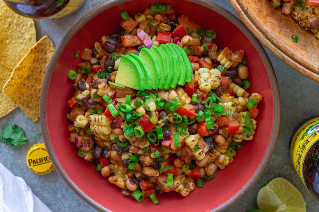 A bowl of colorful cowboy caviar with black beans, corn, diced tomatoes, red bell pepper, and black-eyed peas tossed in a smoky chipotle-lime dressing, garnished with fresh cilantro, served with tortilla chips on the side.