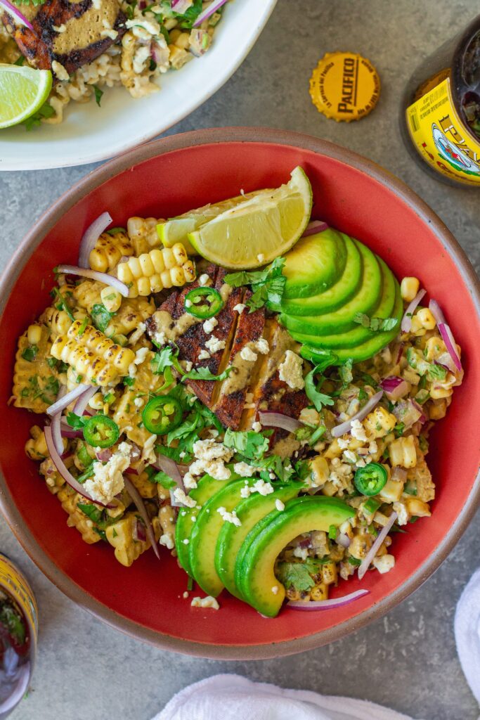 A vibrant street corn tofu rice bowl with crispy golden baked tofu, creamy corn topping, avocado slices, and vegan feta over cilantro lime rice, garnished with lime wedges.