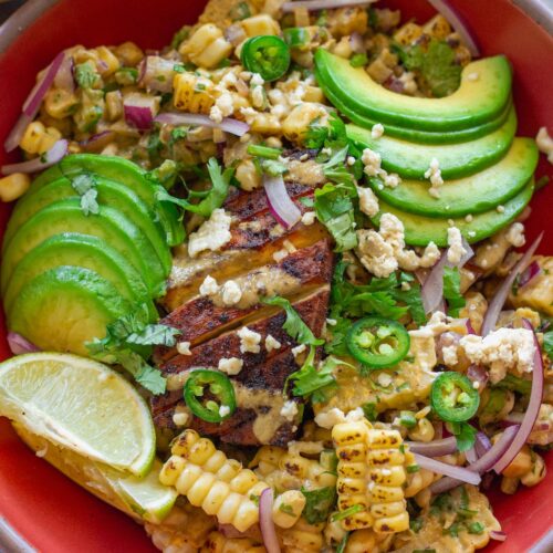 A vibrant street corn tofu rice bowl with crispy golden baked tofu, creamy corn topping, avocado slices, and vegan feta over cilantro lime rice, garnished with lime wedges.