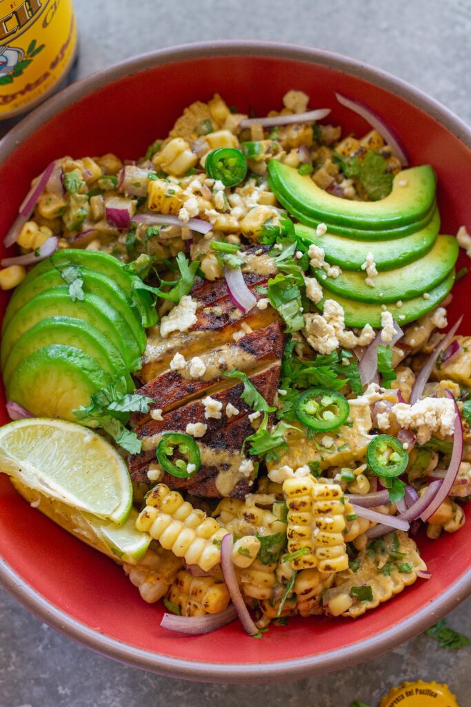 A vibrant street corn tofu rice bowl with crispy golden baked tofu, creamy corn topping, avocado slices, and vegan feta over cilantro lime rice, garnished with lime wedges.