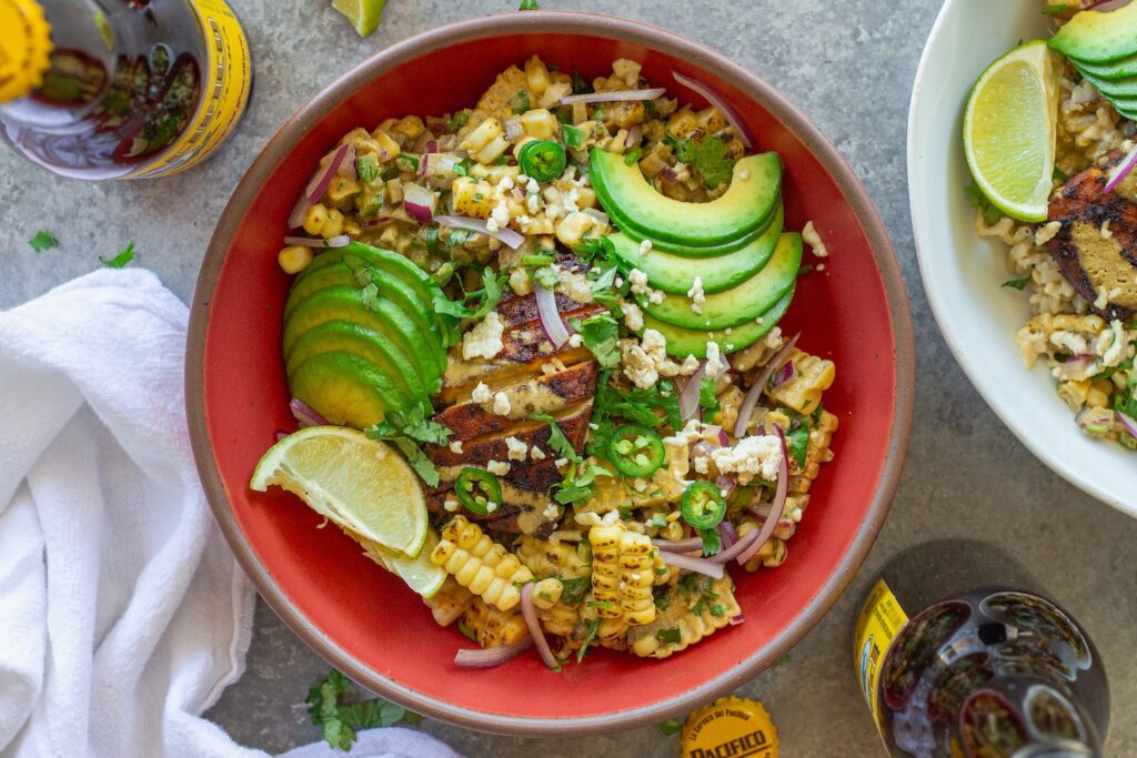 A vibrant street corn tofu rice bowl with crispy golden baked tofu, creamy corn topping, avocado slices, and vegan feta over cilantro lime rice, garnished with lime wedges.