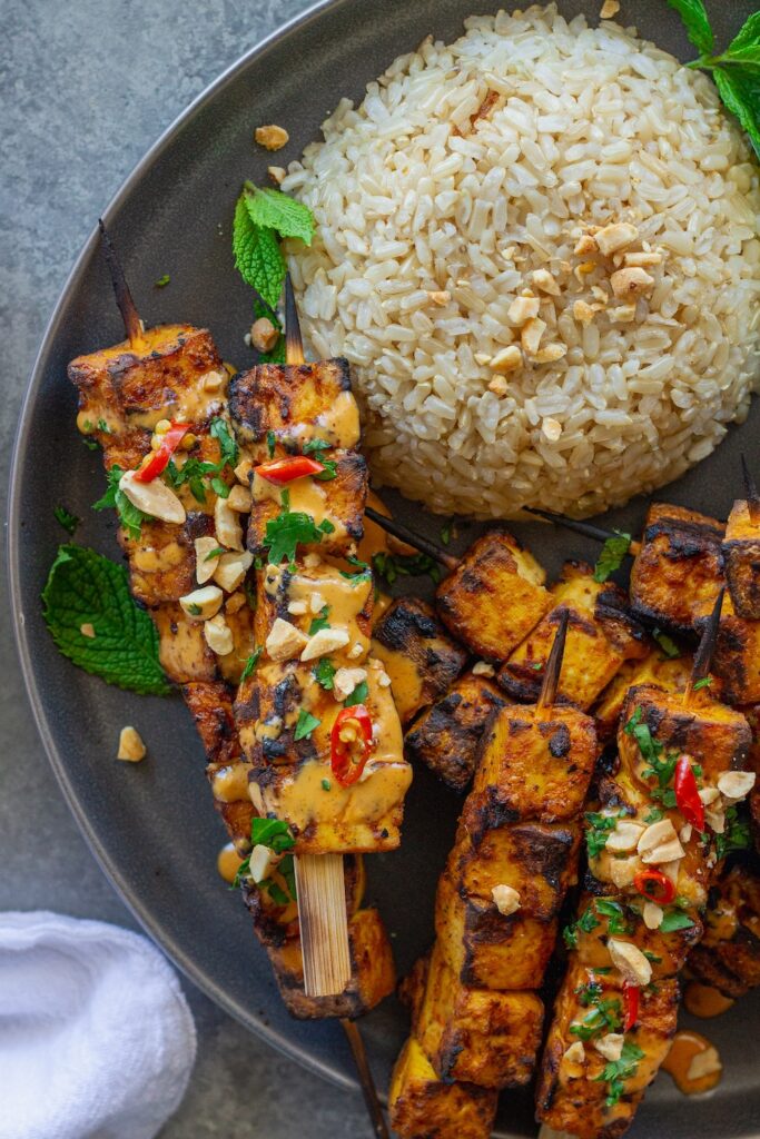 Grilled tofu satay skewers arranged on a platter, drizzled with creamy peanut sauce and garnished with crushed peanuts, fresh cilantro, and Thai red chili slices, served with a side of rice.