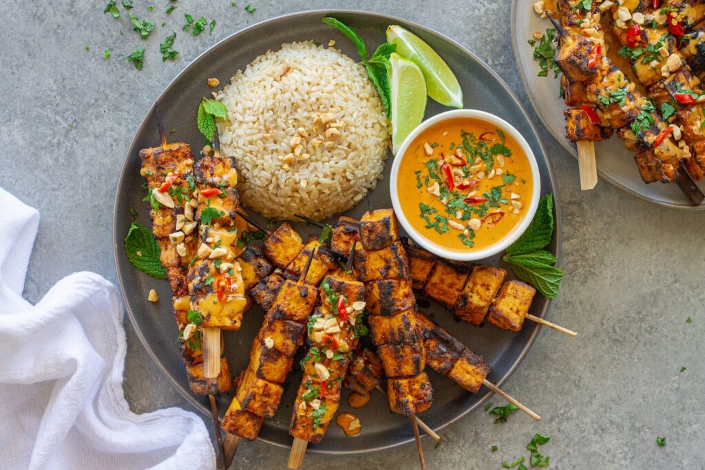 Grilled tofu satay skewers arranged on a platter, drizzled with creamy peanut sauce and garnished with crushed peanuts, fresh cilantro, and Thai red chili slices, served with a side of rice.