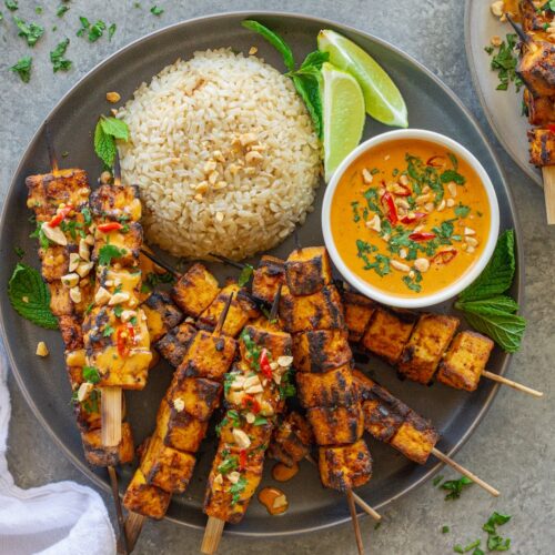 Grilled tofu satay skewers arranged on a platter, drizzled with creamy peanut sauce and garnished with crushed peanuts, fresh cilantro, and Thai red chili slices, served with a side of rice.