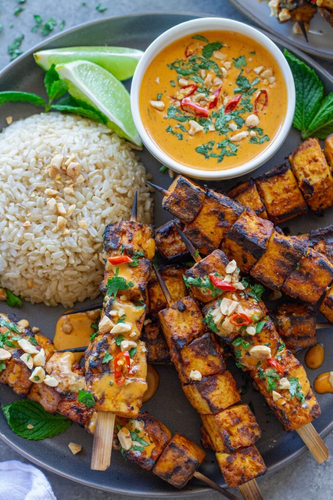 Grilled tofu satay skewers arranged on a platter, drizzled with creamy peanut sauce and garnished with crushed peanuts, fresh cilantro, and Thai red chili slices, served with a side of rice.