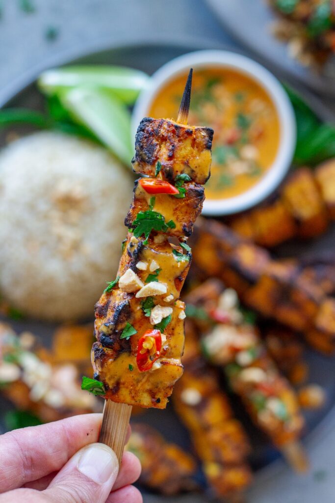 Grilled tofu satay skewers arranged on a platter, drizzled with creamy peanut sauce and garnished with crushed peanuts, fresh cilantro, and Thai red chili slices, served with a side of rice.