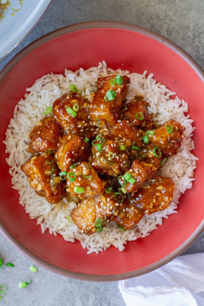 Crispy orange glazed tofu served over white rice, coated in a glossy orange sauce and topped with sliced green onions and toasted sesame seeds.