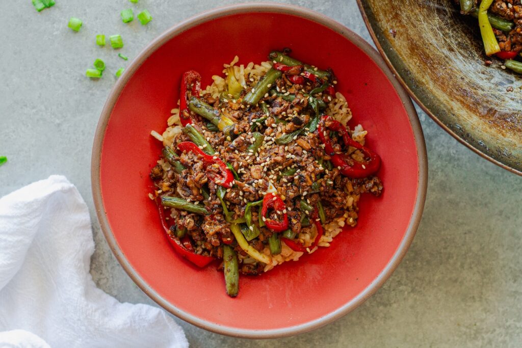 Mongolian tofu stir-fry with crispy grated tofu, green beans, and red chilies served over steamed rice with toasted sesame seeds garnish