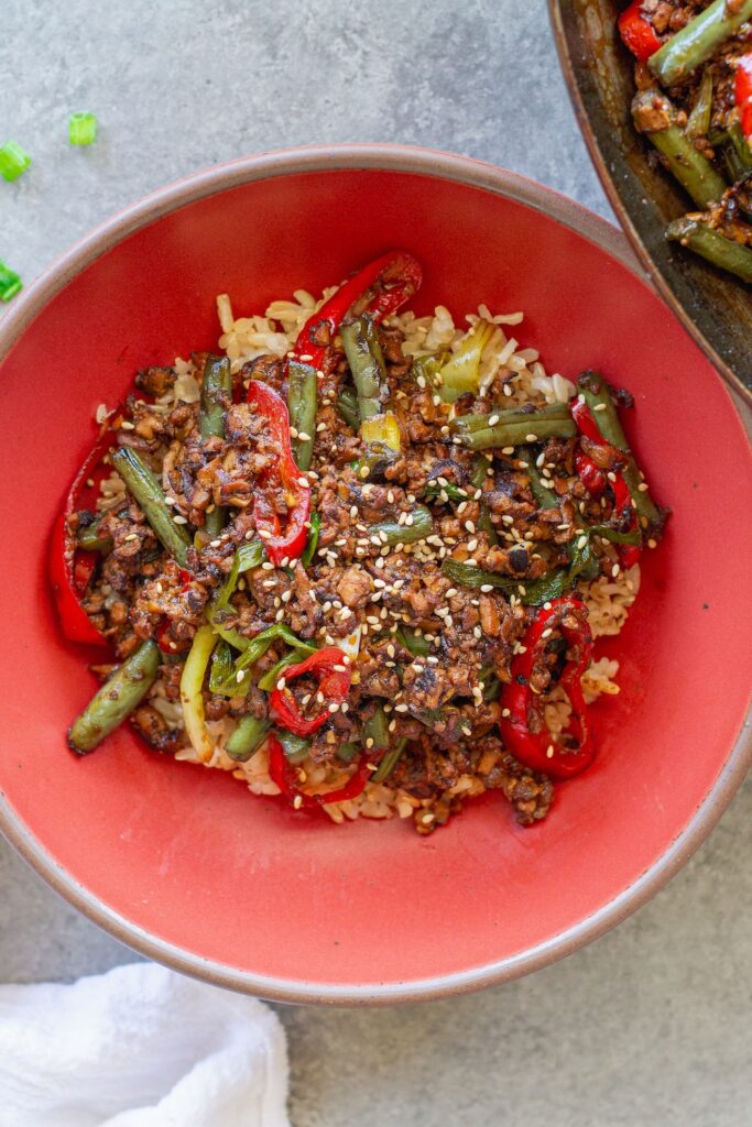 Mongolian tofu stir-fry with crispy grated tofu, green beans, and red chilies served over steamed rice with toasted sesame seeds garnish