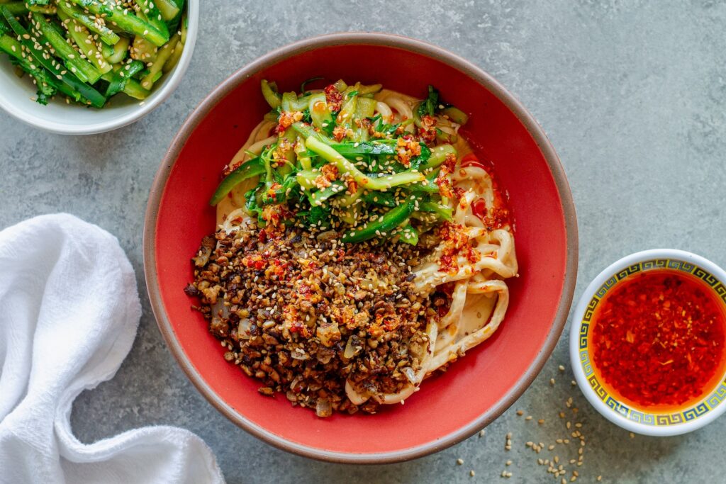 Bowl of chewy udon noodles coated in creamy peanut sauce, topped with crispy shredded tofu, cucumber-cilantro tiger salad, sesame seeds, and a drizzle of chili oil.