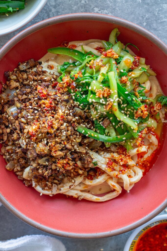 Bowl of chewy udon noodles coated in creamy peanut sauce, topped with crispy shredded tofu, cucumber-cilantro tiger salad, sesame seeds, and a drizzle of chili oil.