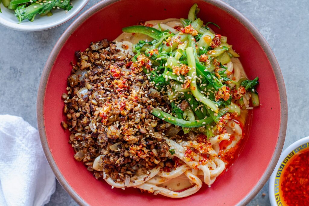 Bowl of chewy udon noodles coated in creamy peanut sauce, topped with crispy shredded tofu, cucumber-cilantro tiger salad, sesame seeds, and a drizzle of chili oil.